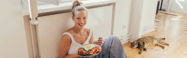 Donna bionda in alto che tiene la colazione sul piatto vicino alla piega scozzese gatto sul pavimento a casa, banner — Foto stock