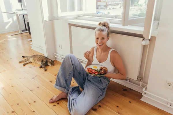Mulher loira feliz segurando café da manhã na placa perto escocês dobra gato em casa — Fotografia de Stock