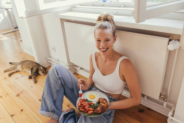 Mujer sonriente en la parte superior del plato de celebración con sabroso desayuno cerca borrosa gato pliegue escocés en casa - foto de stock