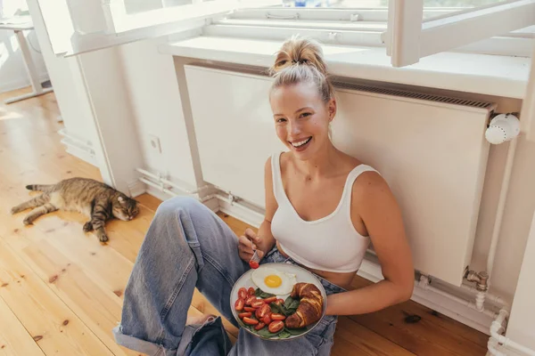 Blonde woman in top holding plate with fried egg and fresh croissant near blurred scottish fold cat at home — Stock Photo