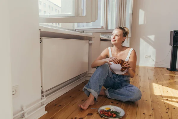 Lächelnde Frau hält Croissant am offenen Fenster und frühstückt zu Hause — Stockfoto