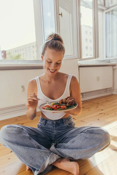 Donna positiva in alto e jeans in possesso di piatto con gustosa colazione sul pavimento a casa — Foto stock