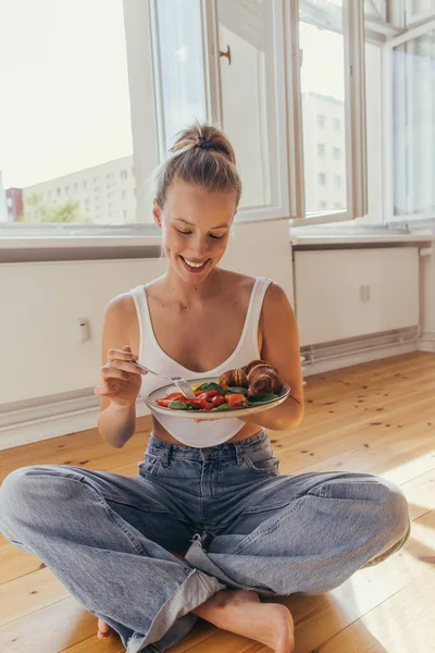 Mujer positiva en la parte superior y vaqueros sosteniendo plato con sabroso desayuno en el suelo en casa - foto de stock