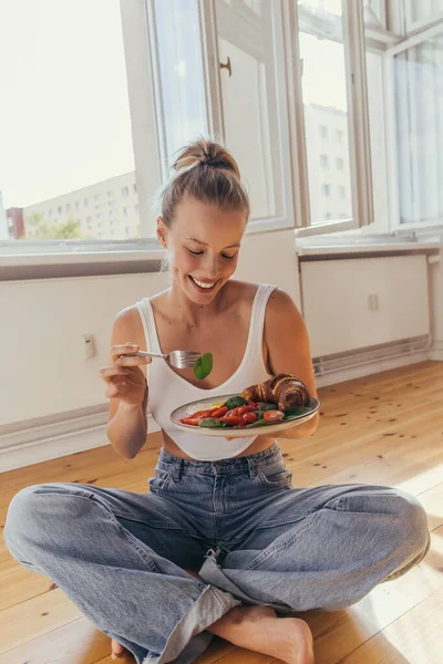 Smiling young woman holding delicious breakfast on plate while sitting on floor at home — Stock Photo