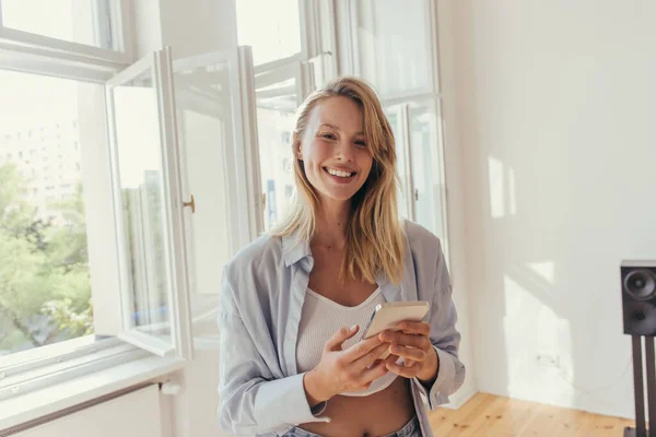 Femme blonde souriante tenant un smartphone et regardant la caméra à la maison — Photo de stock
