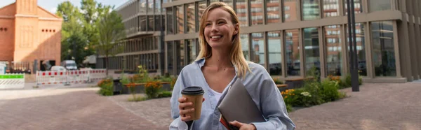 Cheerful blonde freelancer holding paper cup and laptop on urban street in Berlin, banner — Stock Photo
