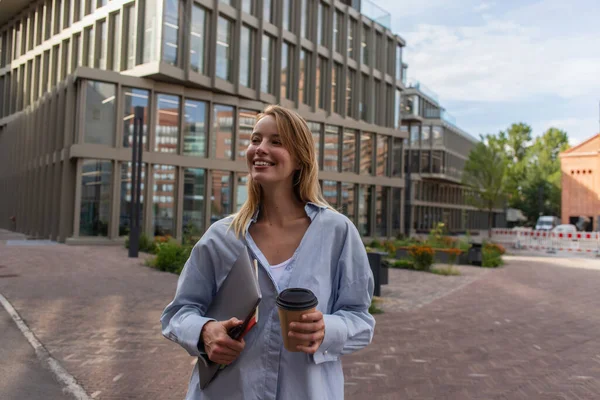 Mujer rubia positiva sosteniendo portátil y bebida para llevar en la calle en Berlín - foto de stock