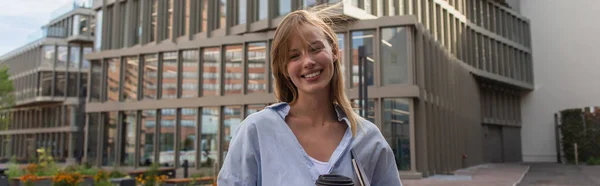Веселая молодая женщина с ноутбуком и кофе, чтобы подойти к размытому зданию на улице в Берлине, баннер — стоковое фото