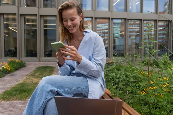 Freelancer sorridente usando smartphone perto de laptop no banco ao ar livre — Fotografia de Stock