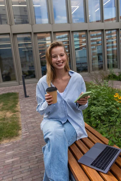 Joyeux pigiste tenant café pour aller et smartphone près d'un ordinateur portable sur le banc à l'extérieur — Photo de stock