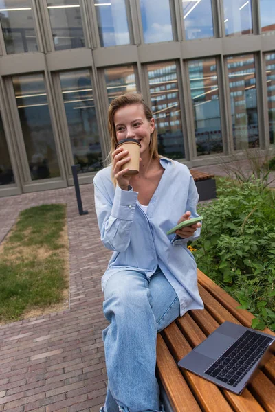 Young woman holding smartphone and drinking coffee to go near laptop on bench on street — Stock Photo