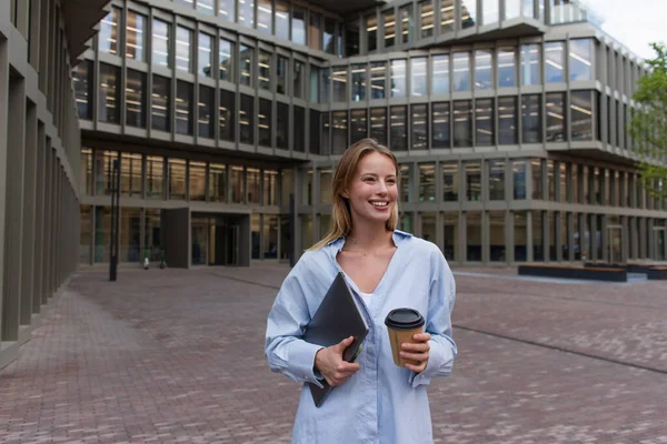 Cheerful young woman holding laptop and paper cup on street in Berlin — Stock Photo