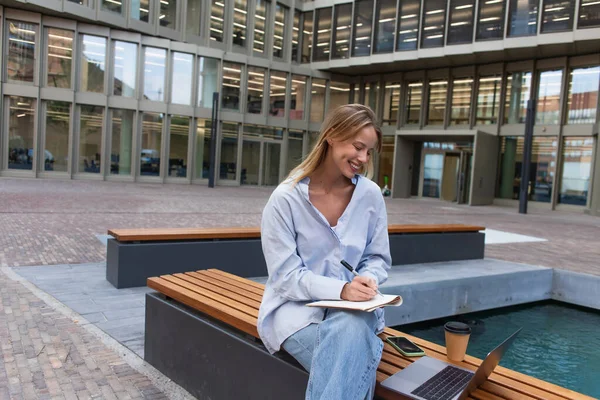 Joyeux jeune femme écrivant sur ordinateur portable près des appareils et du café pour aller sur le banc près de la fontaine à Berlin — Photo de stock