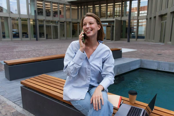 Freelancer sonriente hablando en smartphone cerca del portátil y taza de papel en el banco cerca de la fuente en Berlín - foto de stock