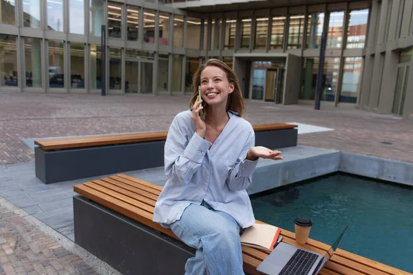 Freelancer hablando por celular cerca del portátil y café en el banco en Berlín - foto de stock