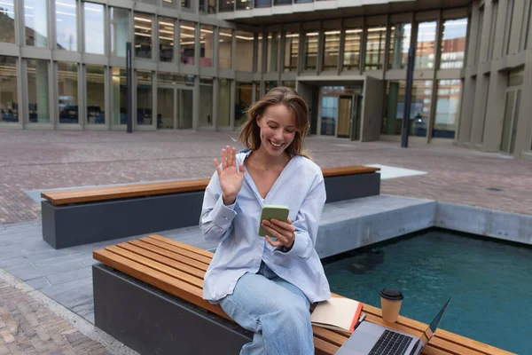 Positive freelancer having video call on smartphone near laptop and paper cup on bench on street in Berlin — Stock Photo