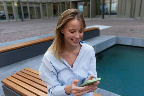 Mujer rubia positiva en camisa usando teléfono móvil en el banco al aire libre - foto de stock