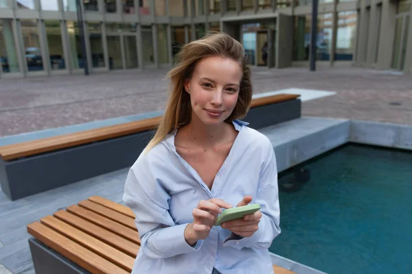 Mujer rubia joven en camisa sosteniendo el teléfono celular y mirando a la cámara cerca de la fuente en la calle urbana - foto de stock