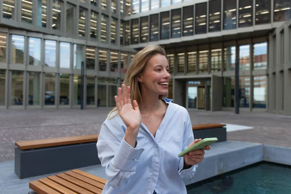 Happy blonde woman in shirt holding smartphone and waving hand on urban street — Stock Photo