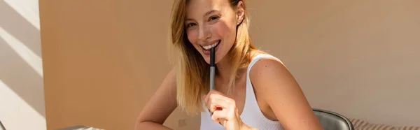 Femme blonde positive tenant un stylo et regardant la caméra à la maison, bannière — Photo de stock