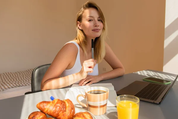 Молодая блондинка держит ручку рядом с ноутбуком и устройствами во время завтрака дома — стоковое фото