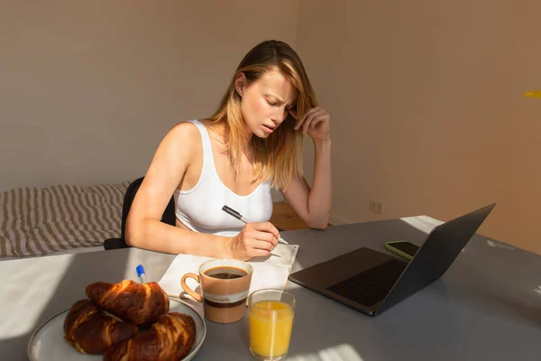 Worried freelancer writing on notebook near devices and breakfast on table at home — Stock Photo