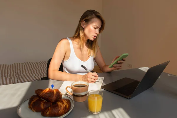 Konzentrierte Freiberuflerin nutzt Smartphone und schreibt am Notebook in der Nähe des Frühstücks zu Hause — Stockfoto