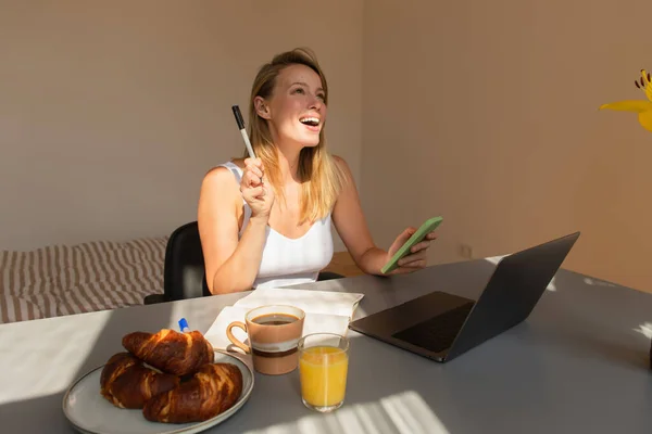 Aufgeregte Freiberuflerin mit Smartphone und Stift in der Nähe von Laptop und Frühstück zu Hause — Stockfoto