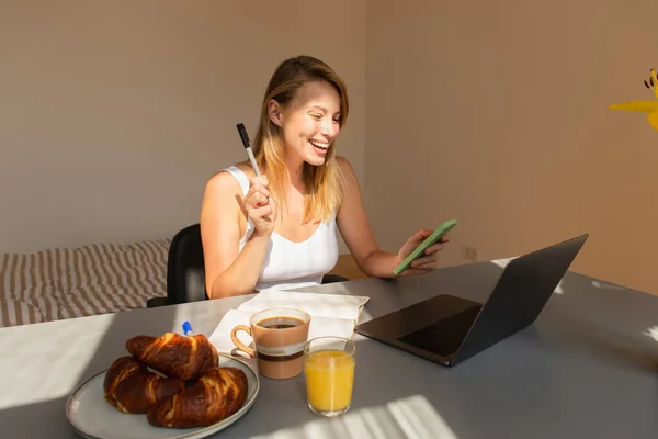 Femme heureuse utilisant un smartphone près d'un ordinateur portable et petit déjeuner à la maison — Photo de stock