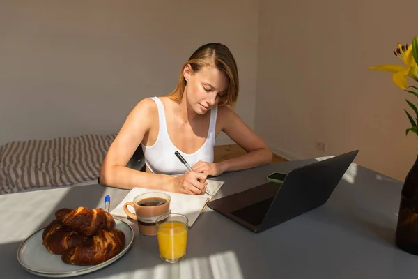 Blonde freelancer in top writing on notebook near devices and breakfast at home in morning — Stock Photo