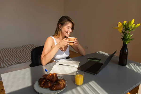 Freelancer sorrindo segurando copo perto do café da manhã e dispositivos em casa — Fotografia de Stock