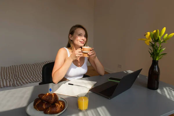 Freelancer satisfecho sosteniendo taza de café cerca de gadgets y croissants en casa - foto de stock