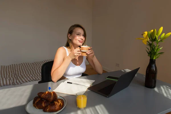 Zufriedene Freiberufler halten Tasse Kaffee in der Nähe von Gadgets und Croissants zu Hause — Stockfoto