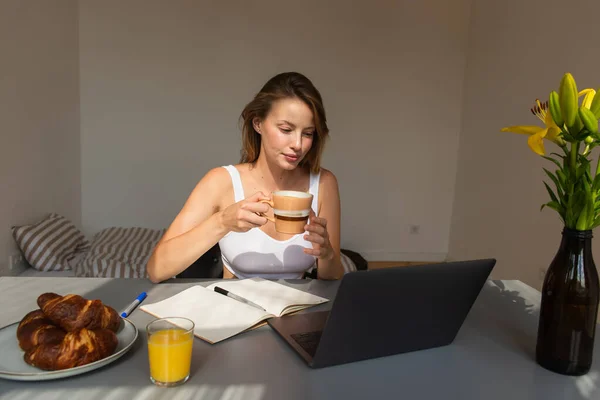 Mulher loira segurando copo e olhando para laptop perto de flores e café da manhã em casa — Fotografia de Stock