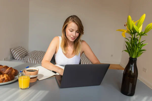 Femme blonde utilisant un ordinateur portable près du petit déjeuner et des boissons à la maison — Photo de stock