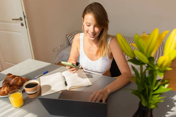 Lächelnde Frau benutzt Geräte in der Nähe von Notebook und Frühstück zu Hause — Stockfoto