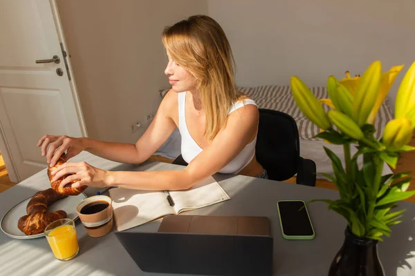 Mujer joven tomando croissant cerca de bebidas y dispositivos en la mesa en casa - foto de stock