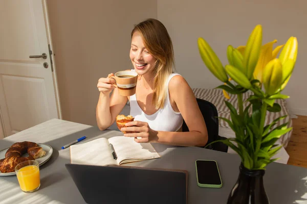 Fröhliche Freiberuflerin mit Kaffee und Croissant in der Nähe von Gadgets und Blumen zu Hause — Stockfoto