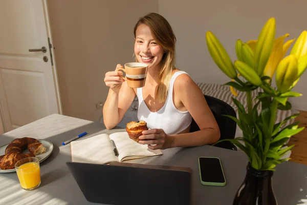 Positive freelancer holding coffee and croissant near devices and flowers at home — Stock Photo