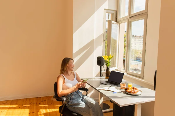 Vista lateral do freelancer sorrindo usando smartphone perto de laptop e café da manhã em casa — Fotografia de Stock