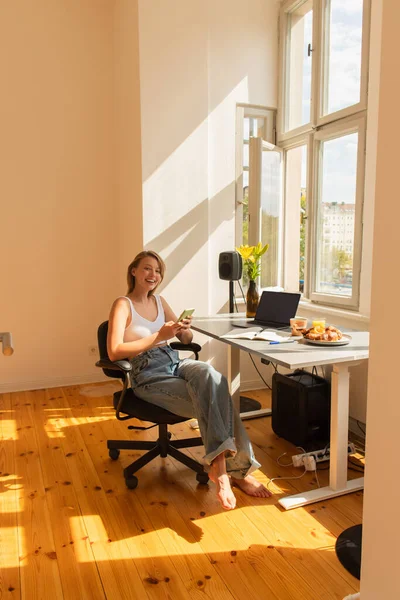 Mulher positiva usando smartphone perto de laptop e café da manhã em casa — Fotografia de Stock