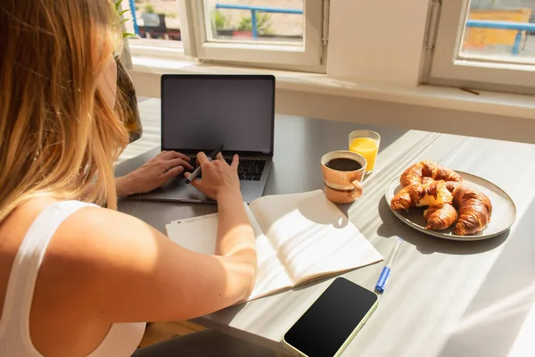 Jovem mulher usando laptop perto notebook e pequeno-almoço na mesa em casa — Fotografia de Stock