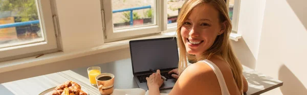 Femme blonde regardant la caméra tout en utilisant un ordinateur portable près du petit déjeuner à la maison, bannière — Photo de stock