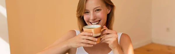 Femme blonde positive tenant une tasse de café et regardant la caméra à la maison, bannière — Photo de stock