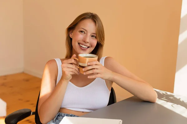 Jovencita alegre en la parte superior sosteniendo la taza de café cerca de la mesa en casa - foto de stock