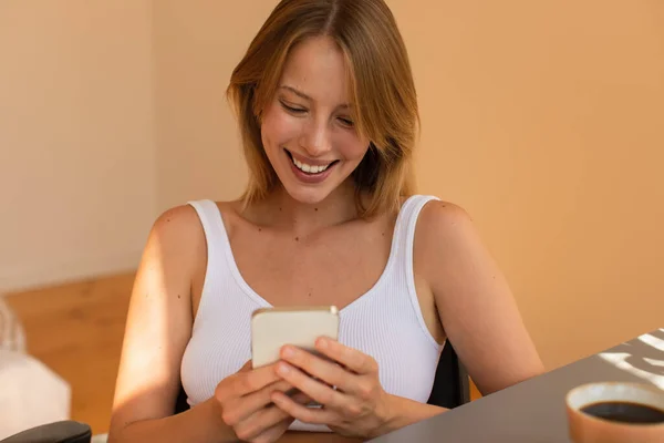 Mulher loira sorridente usando smartphone perto de café borrado em casa — Fotografia de Stock