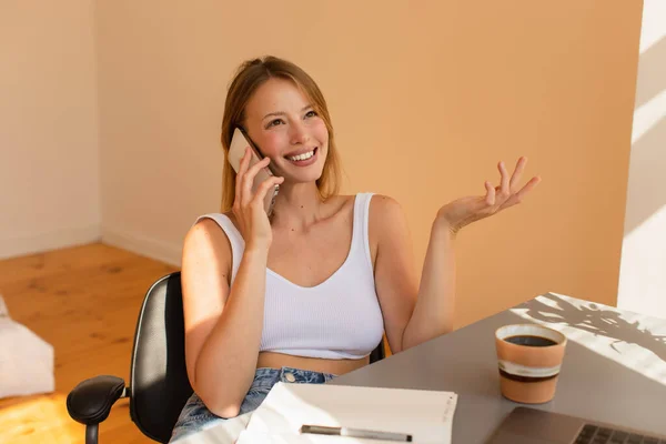 Femme blonde souriante parlant sur smartphone près de café et ordinateur portable à la maison — Photo de stock