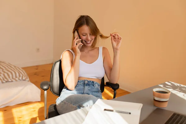 Mujer sonriente tocando el cabello mientras habla en el teléfono inteligente cerca de la computadora portátil y el café en casa - foto de stock