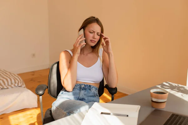 Upset freelancer talking on smartphone near laptop and blurred coffee at home — Stock Photo