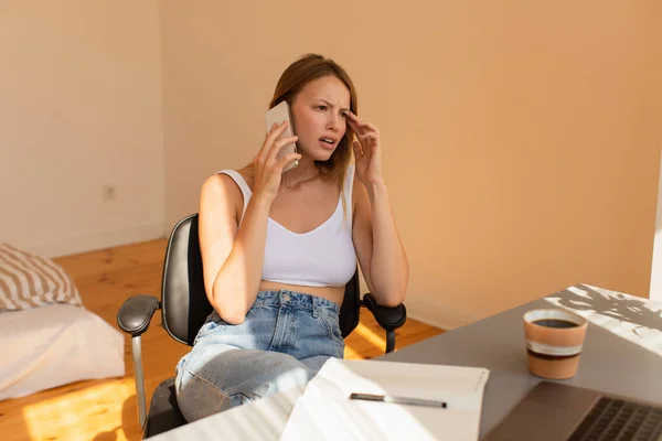 Inquiète freelance parler sur un téléphone mobile près d'un ordinateur portable et une tasse de café à la maison — Photo de stock